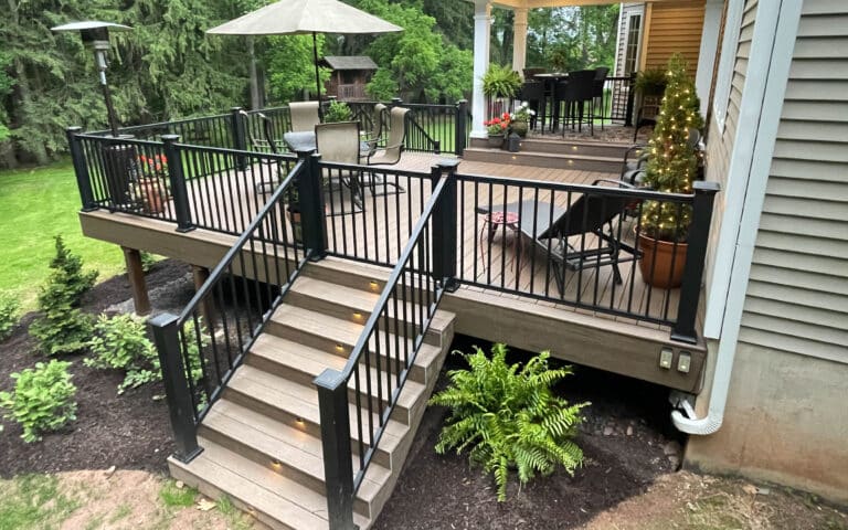A tan 12x20 deck, partly protected by a roof, with seats, an umbrella, lit-up stairs, and has a staircase leading down to the backyard