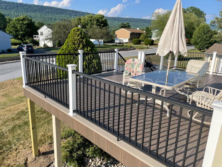 The edge of an elevated 12x20 deck with a black railing, a glass table, seats, and an umbrella