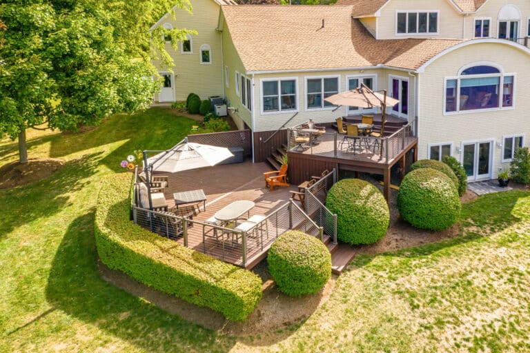 A multi-level 12x20 deck surrounded by bushes and grass, featuring two umbrellas, outside of a tan house with many windows